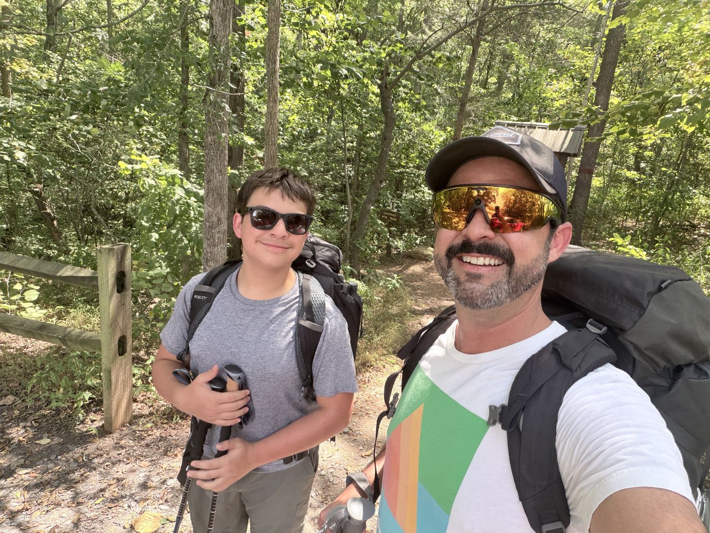 Rob and his son smiling at a trailhead with backpacks and trekking poles.
